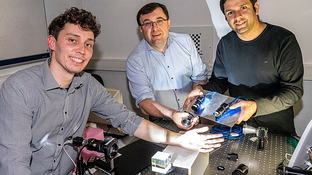 Drei männliche Wissenschaftler stehen um einen optischen Tisch herauf und zeigen die technischen Materialien, die sie für den Hautkrebsscan benutzen. 